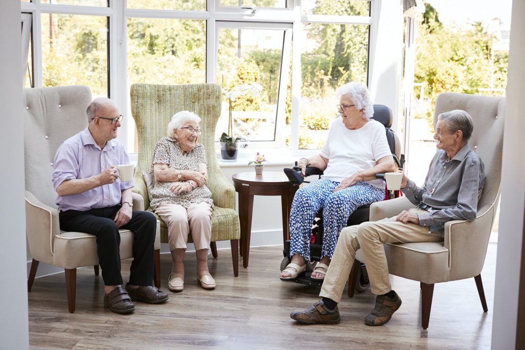 Male And Female Residents Sitting In Chairs And Talking In Lounge Of Retirement Home